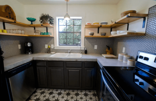 Modern kitchen with gray cabinets, open shelves, a window, and patterned tile flooring. Features coffee maker and sink.