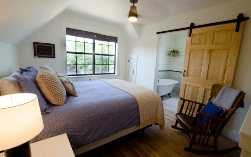 Cozy bedroom with a bed, rocking chair, and a bathroom door in the background, featuring natural light and warm decor.