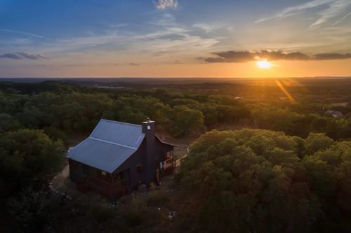 Aerial view of a dark house surrounded by trees at sunset, with vibrant colors in the sky and a peaceful landscape.