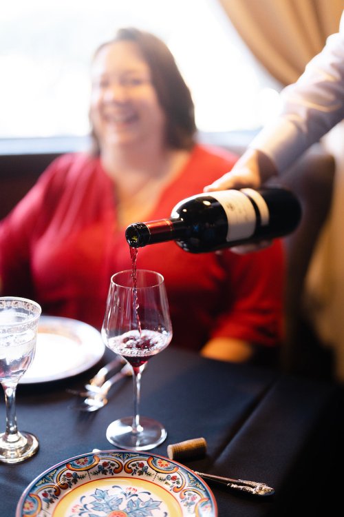 A person pours red wine into a glass at a dining table, with a smiling woman in the background.