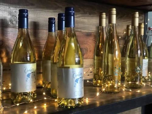 A display of wine bottles on a wooden shelf, illuminated by warm string lights.