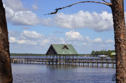A wooden dock extends over calm water, leading to a small, green-roofed pavilion under a blue sky with fluffy clouds.