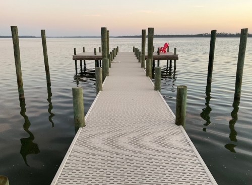 A wooden dock extends over calm water, with a red chair at the end and reflections of the posts in the water.