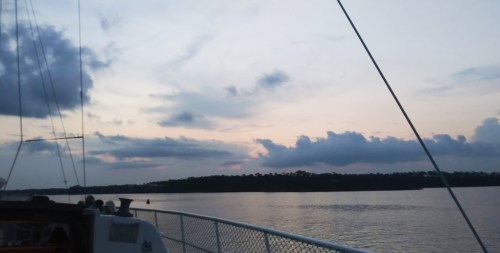 A serene sunset over calm waters, with clouds and silhouettes of trees in the distance, viewed from a boat.