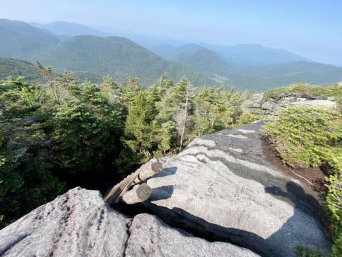 A rocky mountain summit with lush green trees and distant blue mountains under a clear sky.