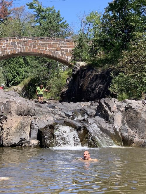 A man swims in a rocky pool beneath a stone bridge, while a person stands on the bank nearby. Lush greenery surrounds the scene.