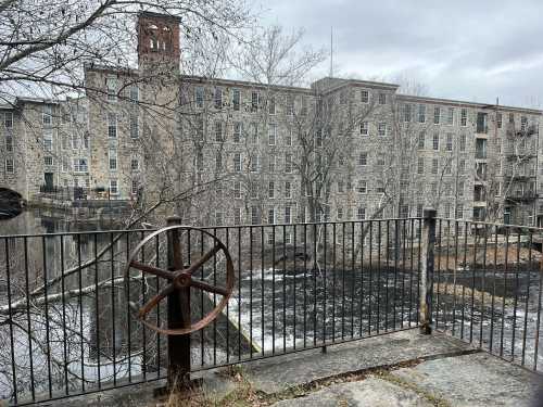 Historic stone mill building beside a river, with a rusted wheel in the foreground and bare trees in a cloudy sky.