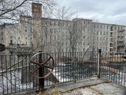 Historic stone mill building beside a river, with a rusted wheel in the foreground and bare trees in a cloudy sky.
