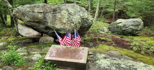 A stone monument with a plaque and three small American flags, surrounded by greenery and large boulders.