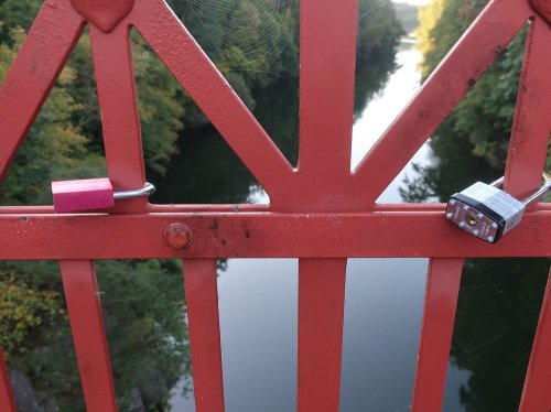 Two padlocks attached to a red metal bridge railing, overlooking a calm river surrounded by trees.