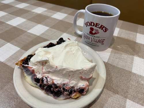 A slice of cherry pie topped with whipped cream, next to a coffee mug from Yoder's Restaurant.