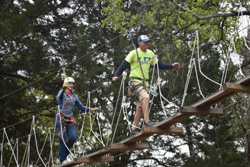 Two people walk across a suspended rope bridge in a forested area, wearing safety gear and harnesses.
