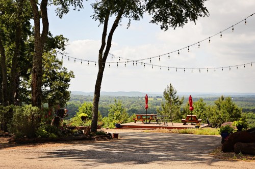 Scenic outdoor area with picnic tables, string lights, and a view of rolling hills under a partly cloudy sky.