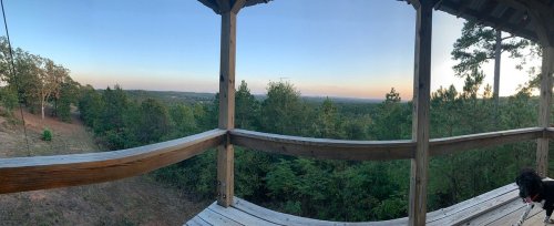 A panoramic view from a wooden deck overlooking a lush green landscape and distant hills at sunset.