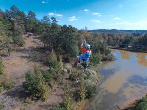 A person zip-lining over a scenic landscape with trees and a pond below, under a clear blue sky.