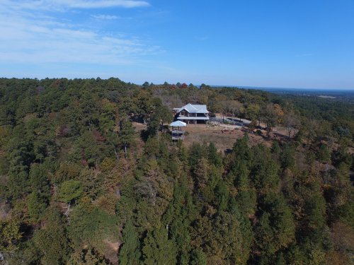 Aerial view of a house surrounded by trees on a hillside, with a clear blue sky in the background.