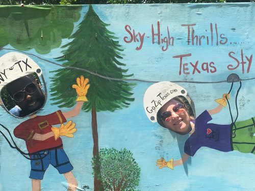 Two people with cut-out faces posing in a colorful mural featuring trees and a zip line theme.