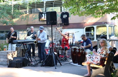 A lively band performs outdoors while a woman relaxes on a bench nearby, surrounded by trees and a vintage vehicle.