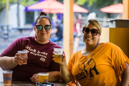 Two women sitting at a table, each holding a cup of beer, smiling and wearing sunglasses at an outdoor event.