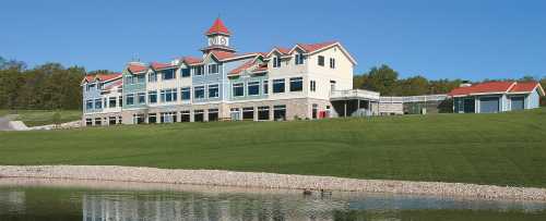 A large, modern building with a clock tower, set on a green lawn by a calm lake under a clear blue sky.