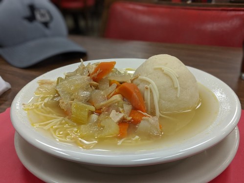A bowl of soup with vegetables, chicken, and a dumpling, served on a table with a gray cap in the background.