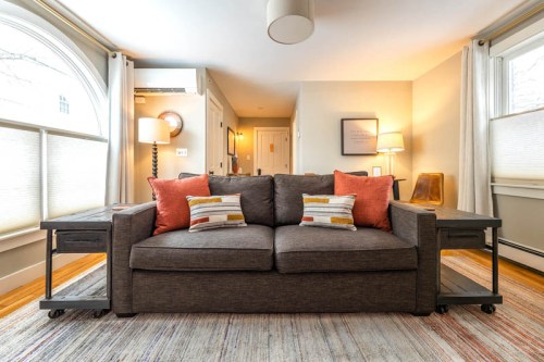 Cozy living room featuring a gray sofa with orange pillows, large windows, and warm lighting.