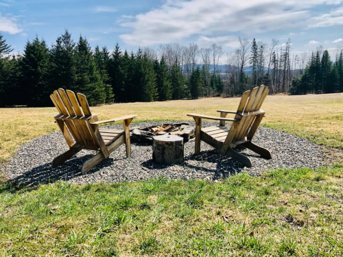 Two wooden chairs facing a fire pit, surrounded by gravel and grass, with trees and a blue sky in the background.