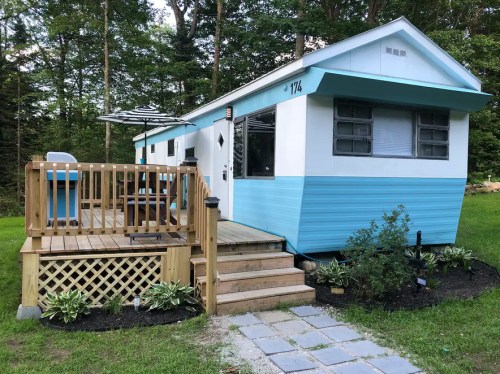 A blue and white mobile home with a wooden deck, steps, and landscaping in a wooded area.
