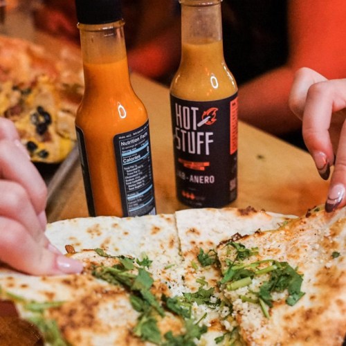 Two hands hold a cheesy quesadilla, with two bottles of hot sauce in the background. Fresh cilantro is sprinkled on top.