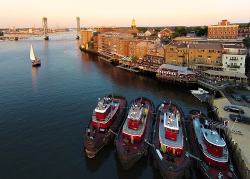 Aerial view of a waterfront with tugboats docked, buildings along the shore, and a sailboat in the distance at sunset.