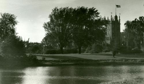 A serene landscape featuring a river, trees, and a historic building with an American flag in the background.