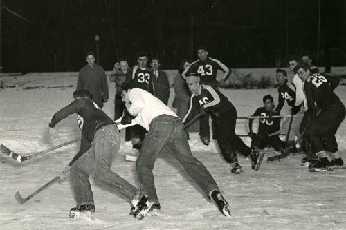 A black-and-white photo of a group of men playing ice hockey on a snowy rink, with players in numbered jerseys.
