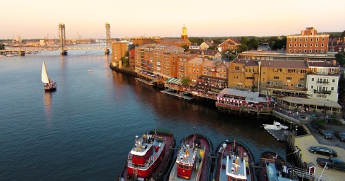 A scenic waterfront view featuring boats, a sailboat, and historic buildings along the shore at sunset.