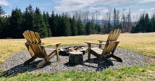Two wooden Adirondack chairs facing a fire pit surrounded by gravel, set in a grassy area with trees in the background.
