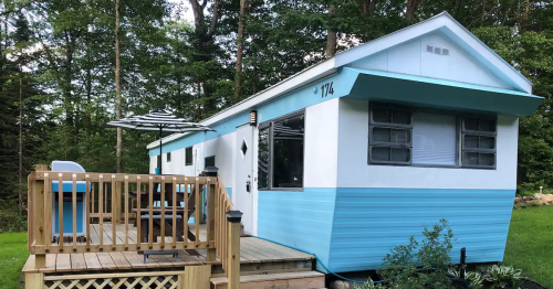 A blue and white mobile home with a wooden deck and patio furniture, surrounded by trees.