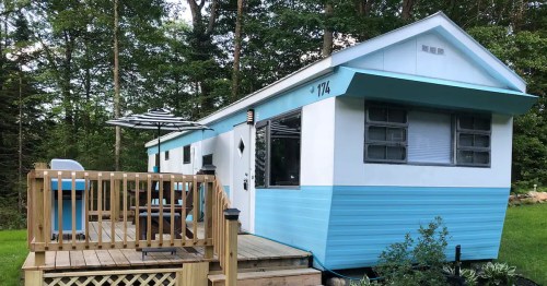 A blue and white mobile home with a wooden deck and patio furniture, surrounded by trees.