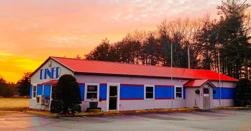 A colorful diner with a red roof and blue stripes, set against a vibrant sunset and surrounded by trees.