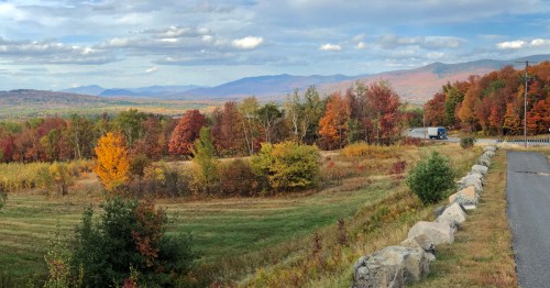 A scenic view of autumn foliage with vibrant orange and yellow trees, mountains in the background, and a winding road.