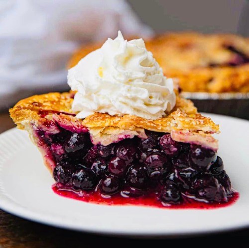 A slice of blueberry pie topped with whipped cream on a white plate, with a whole pie in the background.