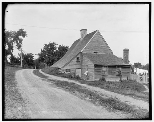 A historic house with a sloped roof, situated on a dirt road, surrounded by trees and laundry hanging outside.