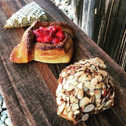 Three pastries on a wooden board: a poppy seed pastry, a fruit-filled croissant, and an almond-covered treat.