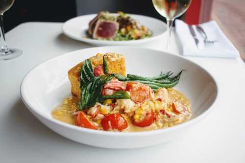 A bowl of creamy risotto with lobster, cherry tomatoes, and greens, served with cornbread, with a glass of wine in the background.