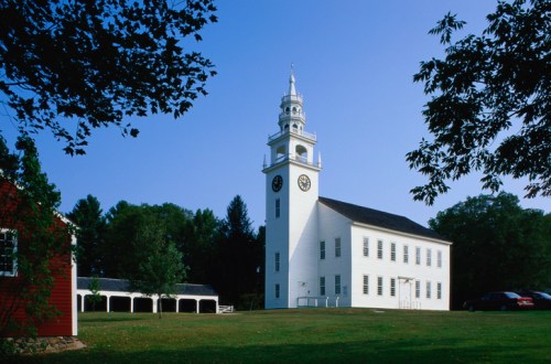 A white church with a tall clock tower, surrounded by green grass and trees under a clear blue sky.