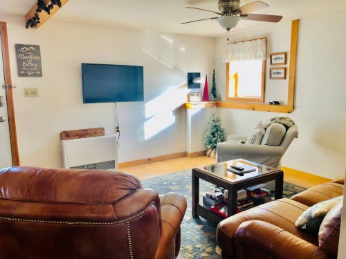 Cozy living room with a TV, comfortable seating, and a small Christmas tree by the window. Warm, inviting atmosphere.