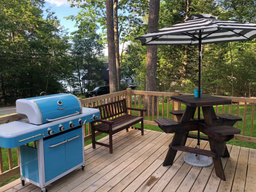 A blue grill next to a picnic table with an umbrella on a wooden deck surrounded by trees and a lake in the background.