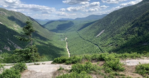 A panoramic view of a lush green valley surrounded by mountains under a partly cloudy sky.