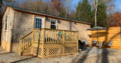 A wooden cabin with a deck, surrounded by trees, featuring outdoor seating and a patio area.