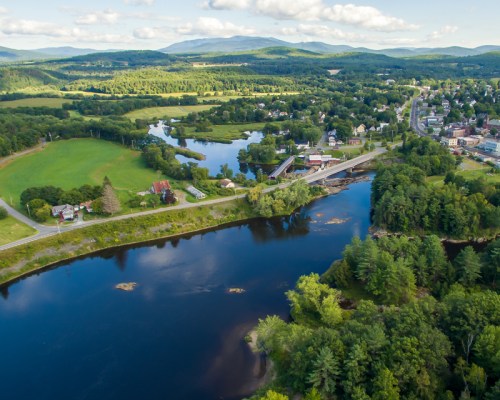 Aerial view of a river winding through green fields and a small town, surrounded by mountains under a blue sky.