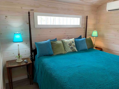 Cozy bedroom with a teal bedspread, decorative pillows, and lamps, featuring wooden walls and a window above the bed.