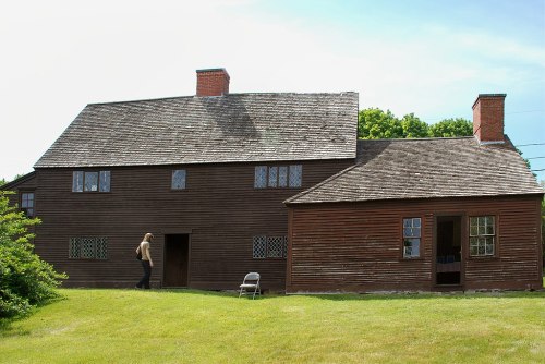 A large, historic wooden house with a sloped roof and a person walking by on a grassy lawn.